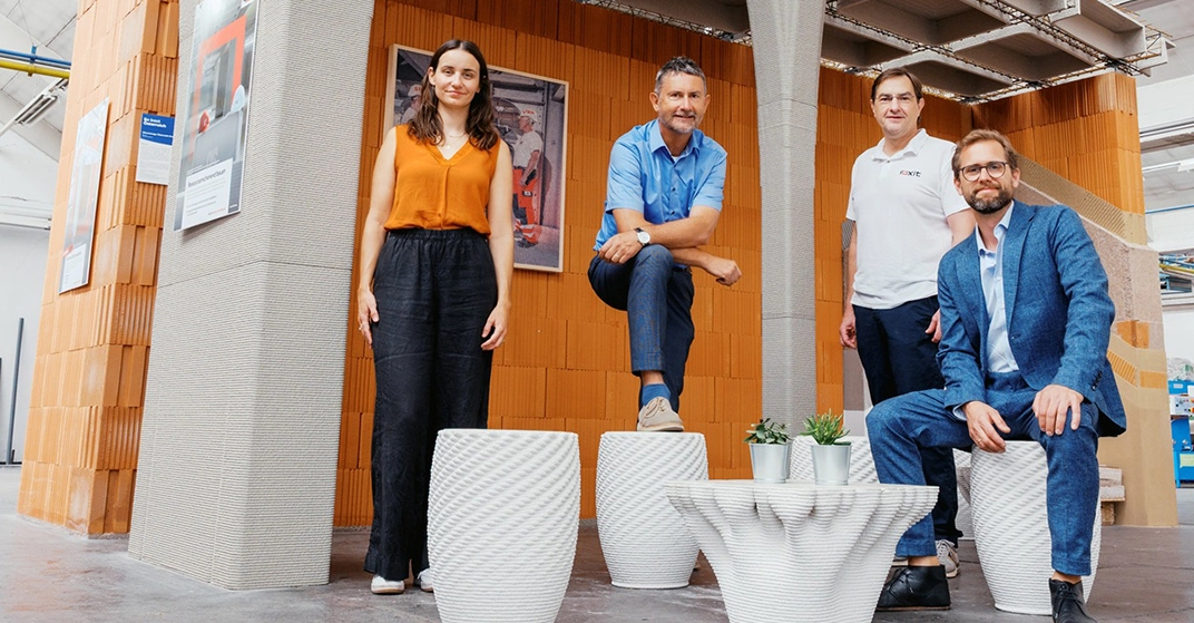 Drei Männer und eine Frau stehen bzw. sitzen auf Gegenständen aus dem 3D-Drucker und lächeln in die Kamera.