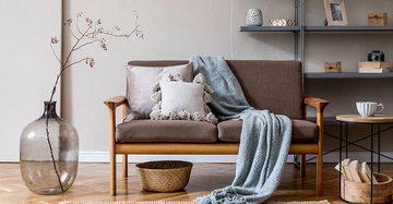 Holzsofa mit Kissen und Decke steht in einem modernen Wohnzimmer neben einer großen Vase mit Zweig Gestelle im Hintergrund halten Dekorationen wie Kerzen und einen Bilderrahmen ein Teppich und ein Tisch ergänzen das Ambiente.