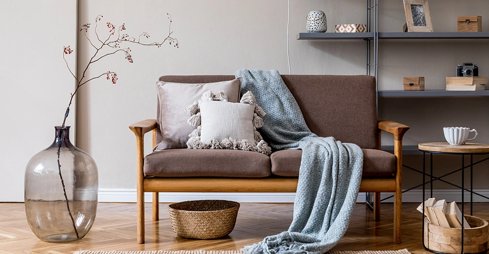 Holzsofa mit Kissen und Decke steht in einem modernen Wohnzimmer neben einer großen Vase mit Zweig Gestelle im Hintergrund halten Dekorationen wie Kerzen und einen Bilderrahmen ein Teppich und ein Tisch ergänzen das Ambiente.