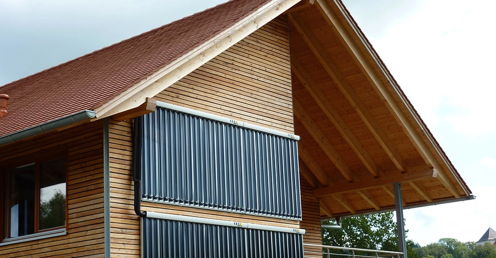 Haus mit schrägem rotem Ziegeldach und Holzfassade montiert auf Balken im Außenbereich zwei Reihen von vertikalen schwarzen Solarpaneelen einige Bäume und bewölkter Himmel im Hintergrund