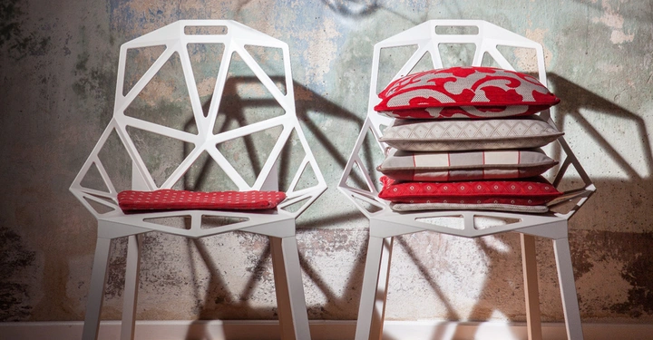 Galerie-Bild 4: Zwei weiße Designerstühle in geometrischem Stil auf Holzboden Eine Vielzahl von gemusterten Kissen auf einem der Stühle verwitterte Wand im Hintergrund