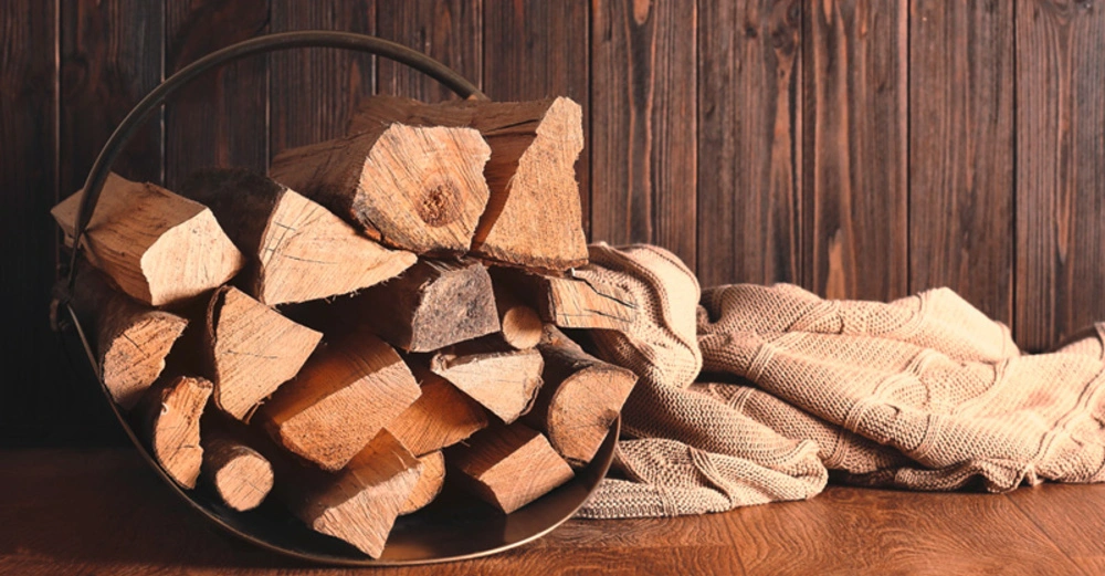 Ein Eimer voller gestapelter Holzscheite steht auf einem Holztisch, daneben liegt eine beige Decke; im Hintergrund eine Holzwand.