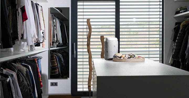 Galerie-Bild 9: Ein begehbarer Kleiderschrank mit Kleidung an beiden Seiten aufgehängt steht im Raum. In der Mitte steht ein Tisch mit Dekorationsgegenständen. Im Hintergrund befindet sich ein großes Fenster mit Jalousien.