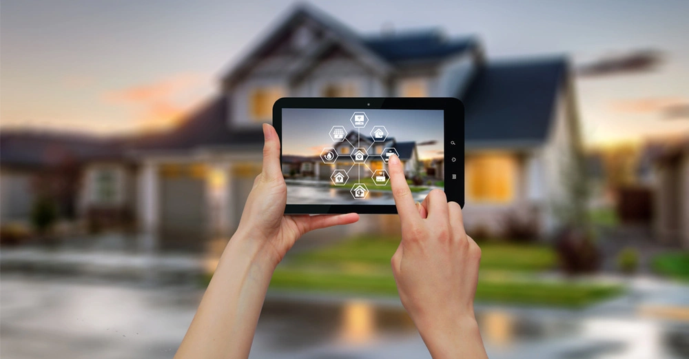 Person bedient ein Smart Home mit Anwesenheitssimulation mit einem Tablet vor dem Haus, das von der Abenddämmerung beleuchtet ist.