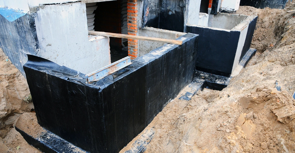 Betonfundament-segment wird bearbeitet in einer Baugrube mit Sandboden. Ein Wasserwaage liegt auf dem Fundament um Ebenheit zu überprüfen. Ziegel und Schalungsbretter sind sichtbar im Aufbau.