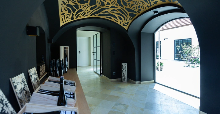 Galerie-Bild 15: Weinflaschen stehen auf einem Holzregal. Sonnenlicht scheint durch große Fensterbogen in einen eleganten Raum mit dunklen Wänden und goldener Deckenverzierung. Dahinter ist ein heller Innenhof sichtbar.