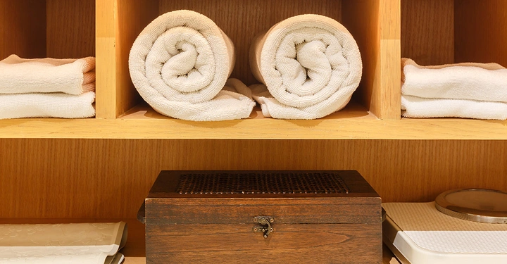 Gerollte und gefaltete weiße Handtücher liegen in einem Regal aus hellem Holz, darunter eine braune Holzkiste und eine Waage.