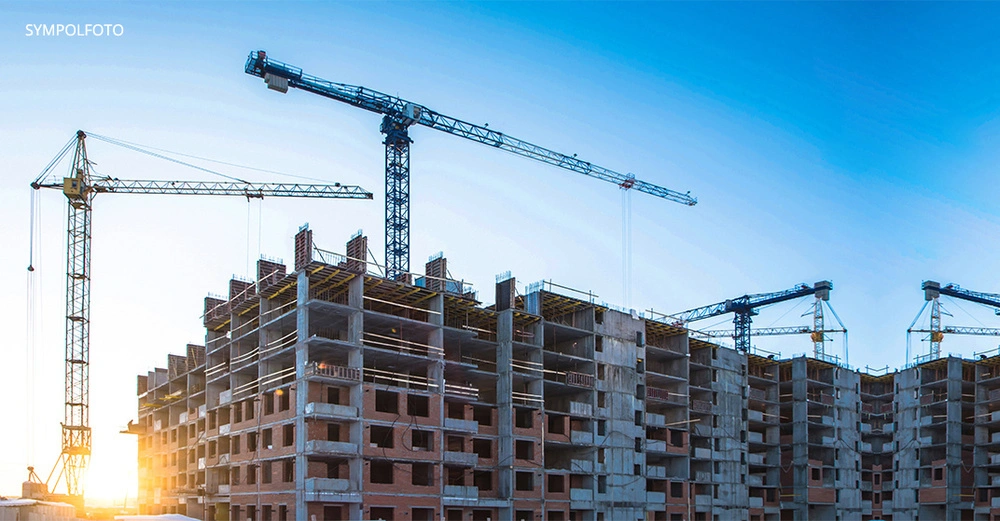 Ein teilweise gebautes Gebäude steht im Vordergrund Bauelemente und Gerüste sind sichtbar Kräne arbeiten am Bau im Hintergrund der Himmel ist klar und bläulich Text: "SYMBOLFOTO"