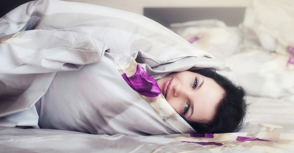 Eine Person liegt gemütlich in einer Decke eingewickelt auf einem Bett mit weiß-violetter Bettwäsche. Die Umgebung ist ein Schlafzimmer mit weiteren Kissen und Decken im Hintergrund.