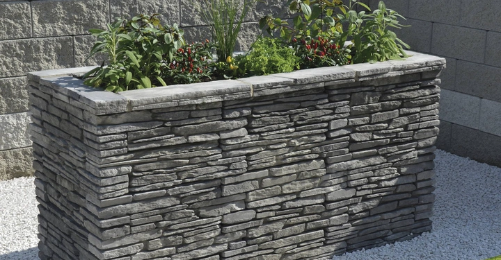 Galerie-Bild 11: Rechteckiger Blumenkasten aus grauem Stein mit grünen Pflanzen und bunten Blumen auf einem Untergrund aus weißen Kieseln vor einer grauen Steinmauer.