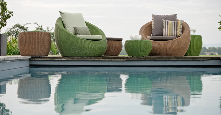 Galerie-Bild 7: Zwei runde, geflochtene Stühle mit Kissen, umgeben von passenden Beistelltischen, stehen nebeneinander auf einer Terrasse, vor einem Pool, mit Pflanzen im Hintergrund.