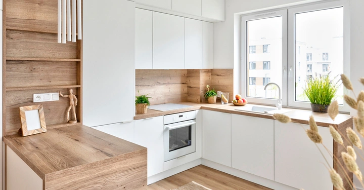 moderne, sehr helle Küche mit Holzvertäfelung und Tisch in Holz, Kücheneinrichtung mit weißen Fronten, rechts ein großes Fenster mit Pflanzen davor.