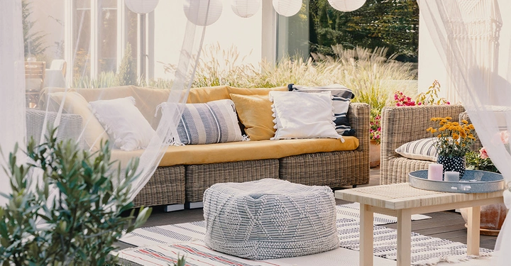 Rattan-Sofa mit gelben Kissen steht auf einer Terrasse neben einem kleinen Tisch mit Blumen und Kerzen umgeben von Pflanzen und dekorativen Lichtern im Hintergrund.