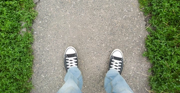 Fußgänger in heller Jeans und schwarzen Converse steht auf schmalem, asphaltiertem Gehweg, rechts und links davon Wiese