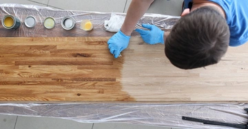 Eine Person trägt Holzlack auf eine lange Holzplatte auf. Sie trägt blaue Handschuhe. Daneben stehen mehrere Farbdosen auf einer Plastikfolie auf einem Tisch.