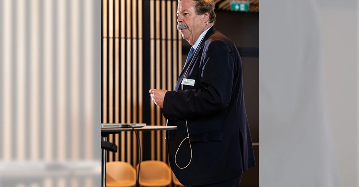 Galerie-Bild 6: Ein Mann in Geschäftskleidung steht vor einer Präsentation in einem modernen Konferenzraum mit Holzverkleidung. Er hält ein Mikrofon und schaut in Richtung des Publikums.