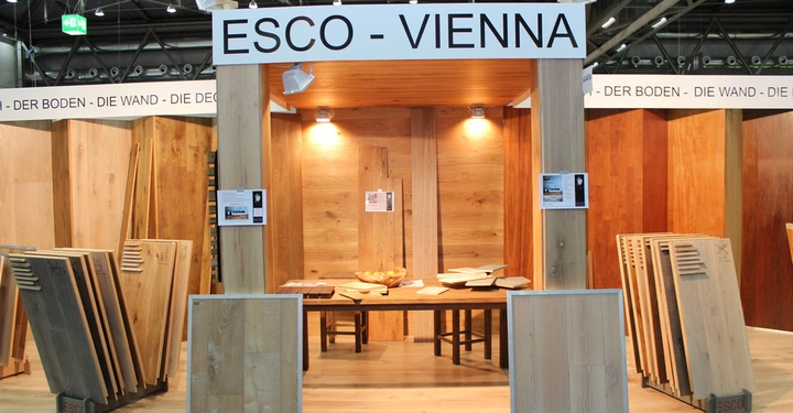 Galerie-Bild 19: Holztafeln stehen gestapelt vor einem Messestand namens ESCO - VIENNA untergebracht in einer großen Halle mit hölzernen Mustern an den Wänden und Holzproben auf einem Tisch.