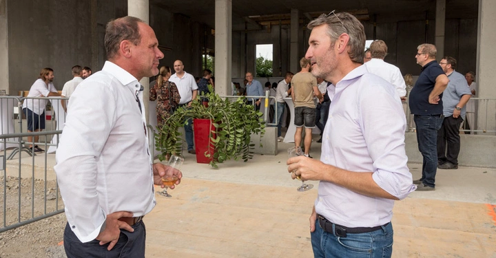 Galerie-Bild 4: Zwei Männer halten Weingläser und unterhalten sich stehend. Im Hintergrund stehen weitere Personen in einer offenen Betonstruktur mit Säulen und Dekorationen an einem Unscharf im HintergrundPartyähnlichem.