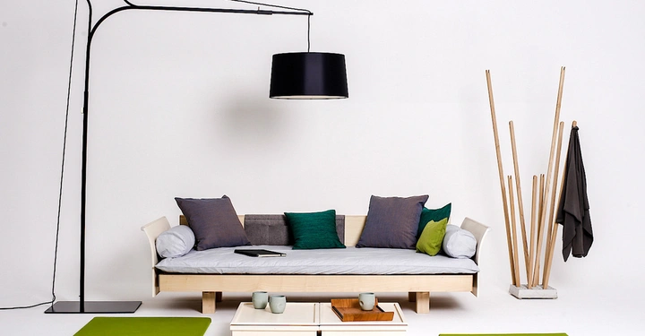 Galerie-Bild 3: Helles Wohnzimmer mit einem weißen Sofa das mit bunten Kissen dekoriert ist Ein schwarzer Lampenschirm hängt darüber Ein Beistelltisch mit Tassen und einem Tablett steht davor Grüne Sitzpolster am Boden rechts ein Kleiderständer und Mantelstab