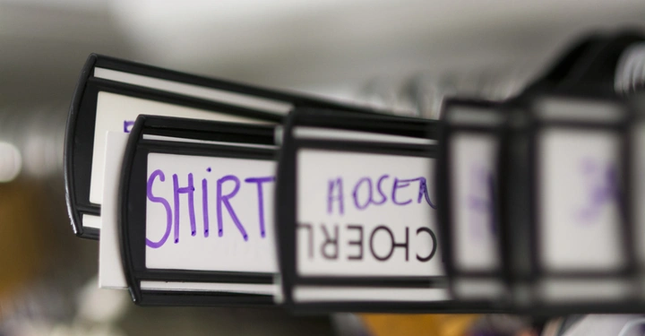 Galerie-Bild 8: Kleiderbügel hängen an einer Stange in einem Geschäft. Auf den Schildern, die an den Bügeln befestigt sind, steht das Wort SHIRT in lila Schrift. Im Hintergrund sind weitere Kleider unscharf sichtbar.