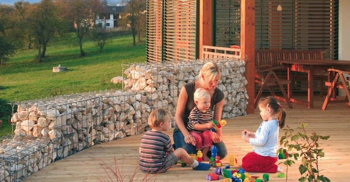 Frau spielt mit drei kleinen Kindern mit bunten Bauklötzen auf einer Holzterrasse umgeben von dekorativen Steinwänden mit grünem Feld und Häusern im Hintergrund