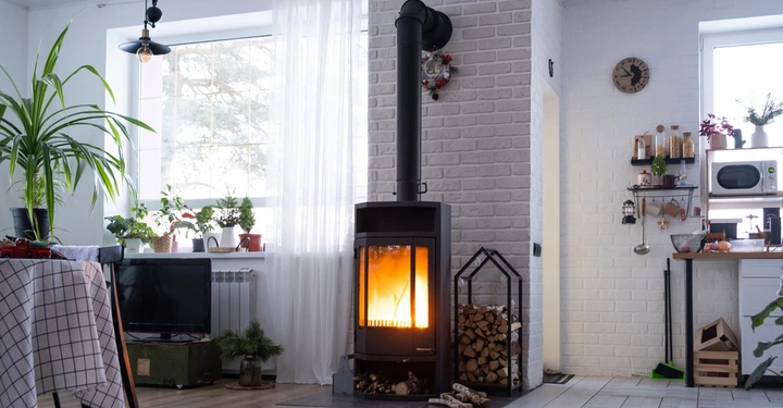 Holzofen brennt in einem gemütlichen Wohnzimmer bei Tageslicht Ofen steht vor einer weißen Backsteinwand Raum dekoriert mit Pflanzen ein Tisch mit kariertem Tuch links und Küchenutensilien rechts.