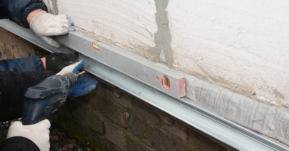 2 Bauarbeiter in Handschuhen dichten freigelegtes Fundament mit Steinbohrer und Metallschiene ab. Zweiter Arbeiter hält Wasserwaage an die Leiste. 