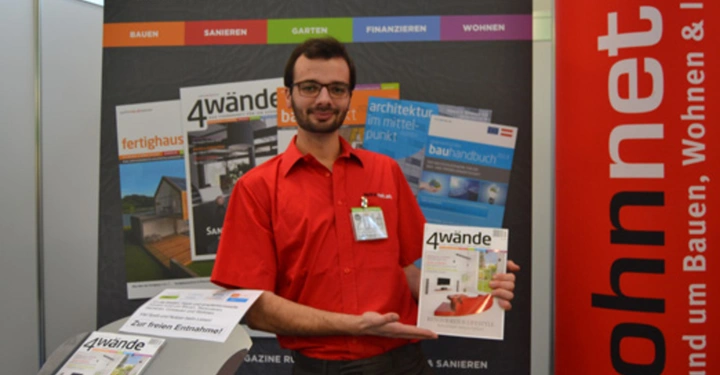 Galerie-Bild 1: Ein Mann hält eine Zeitschrift 4wände in einem Ausstellungsbereich Präsentiert von wohnnet.at mit verschiedenen Werbepostern und einem Stehpult Text auf der Zeitschrift 4wände.