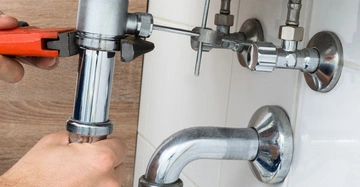 Hände verwenden eine Rohrzange um ein silbernes Rohr unter einem Waschbecken zu reparieren. Im Hintergrund sind Wandfliesen und andere Rohrleitungen sichtbar.