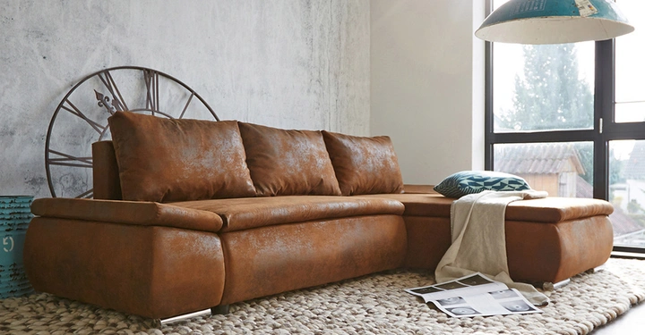 Braunes Ledersofa steht in einem hellen Raum vor einer Wand mit Uhrendekor Ein übergroßes Fenster zeigt Außenansicht Ein blauer Kissen und ein weißes Tuch liegen auf dem Sofa Zeitungen liegen auf einem geflochtenen Teppich