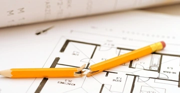 Gebrochener gelber Bleistift liegt auf einem Architekturplan in einem Büro mit Rollen von Bauzeichnungen im Hintergrund.