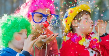 3 Kinder mit bunten Perücken in giftgrün und pink sowie ein gelber Hut und lustige Brille pusten Konfetti in die Luft und blasen in Luftschlangen. Fasching!