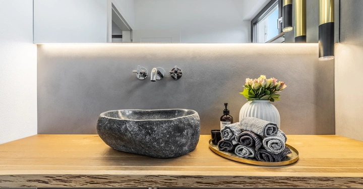 Galerie-Bild 5: Waschbecken steht auf hölzerner Arbeitsplatte enthält Seifenspender und Tablett mit gerollten Handtüchern in modern eingerichtetes Badezimmer mit grau beleuchteten Wänden und Vase mit Blumen. Text: LAMURISTA