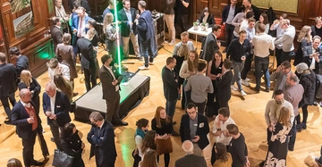 Menschenmenge von oben bei einer Veranstaltung, einige tragen Mund-Nasenschutz, unterhalten sich miteinander, einige mit Trinkglas in der Hand
