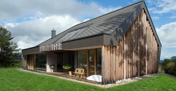 Haus in modernem Stil mit Holzverkleidung steht auf einer grünen Wiese umgeben von Bäumen und Himmel mit Wolken im Hintergrund. Holzstühle und Topfpflanzen stehen auf der Terrasse.