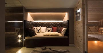 Gemütliche Sitznische mit Kissen und Decken, daneben ein Holzsaunaraum mit Glastür. Der Bereich ist umgeben von dunklem Tapetenmuster und Holzverkleidung, Teppich auf Boden.