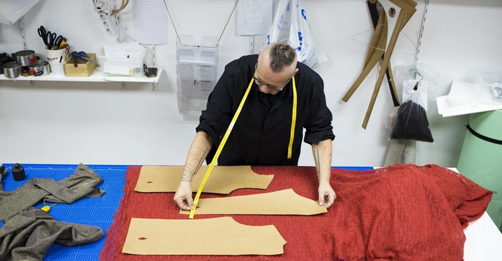 Galerie-Bild 6: Eine Person misst mit einem Maßband Schnittmuster auf rotem Stoff an einem Arbeitstisch ab in einem Nähatelier mit Regalen und Werkzeugen an der Wand.