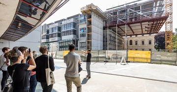 Eine keine Menschengruppe steht vor der sich im Bau befindlichen Unibibliothek Graz, eine junge Frau fotografiert das Gebäude mit dem Handy