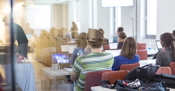Menschen sitzen an Tischen und arbeiten an Laptops in einer hellen Büroumgebung. Einige stehen im Hintergrund und unterhalten sich. Angeschlossene Geräte und Notizen verteilen sich auf den Tischen.