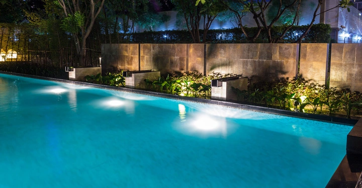 Beleuchteter Swimmingpool bei Nacht in einem modernen Gartenbereich mit Pflanzen und hoch aufragender Steinmauer im Hintergrund der von Bäumen und Buschwerk umgeben ist.