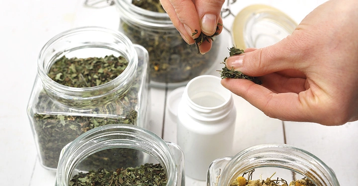 Hände füllen getrocknete Kräuter aus Glasbehältern in eine weiße Plastikflasche in einer hellen Umgebung mit mehreren Kräutergläsern auf einem weißen Tisch