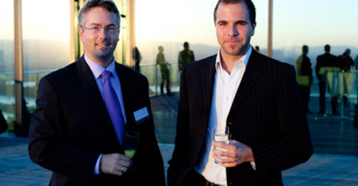 Galerie-Bild 10: Zwei Männer in Anzügen stehen und halten Getränke in der Hand vor einem modernen Gebäude mit Glaswänden. Im Hintergrund sind Personen und eine Abendlandschaft erkennbar.