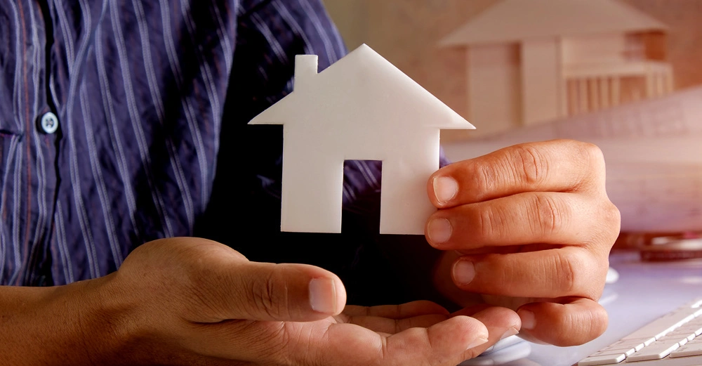 Eine Hand hält ein kleines weißes Modell eines Hauses vor einem Mann in einem gestreiften Hemd sitzend an einem Schreibtisch mit einem Computer im Hintergrund.