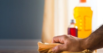 Eine Hand wischt mit einem gelben Tuch eine Oberfläche ab zwei Reinigungsmittelflaschen sind im Hintergrund in einem Wohnraum zu sehen.