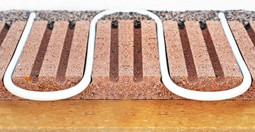 Weiße Rohre schlängeln sich durch gerillte braune Styroporplatten zur Fußbodenheizung installiert auf einer gelben Bodenfläche.