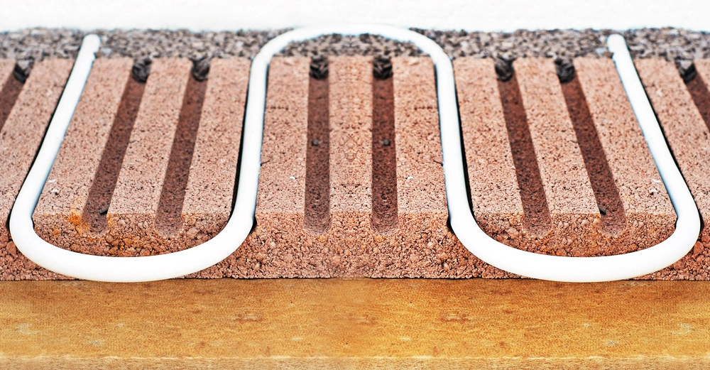 Weiße Rohre schlängeln sich durch gerillte braune Styroporplatten zur Fußbodenheizung installiert auf einer gelben Bodenfläche.