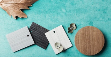 Drei Farbproben und ein runder Holzuntersetzer liegen auf einem grünen Stoff Untergrund. Links oben befindet sich ein braunes getrocknetes Blatt, während oben rechts zwei Blumenblätter sichtbar sind.