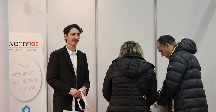 Galerie-Bild 15: Ein Mann im Anzug hält Werbetaschen während sich zwei Personen in Winterjacken etwas an einem Stand ansehen in einem Messeumfeld. Text: wohnnet WO UND WIE WOHNEN Deine Wohnraumverwirklichungsmaschine IMMOBILIEN.