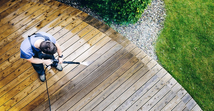 Person reinigt mit einem Hochdruckreiniger eine Holzterrasse im Gartenbereich umgeben von Gras und Kieselsteinen Bepflanzungen am Rand.