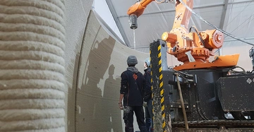 großer 3D-Betondrucker bei der Arbeit. Es entsteht eine hohe, gerillte Betonwand. Der Drucker ist orange, vor ihm steht ein Arbeiter und überwacht das Ganze.